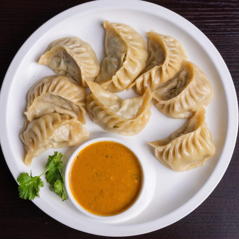 A plate of steamed momos with a side of orange dipping sauce and fresh cilantro.