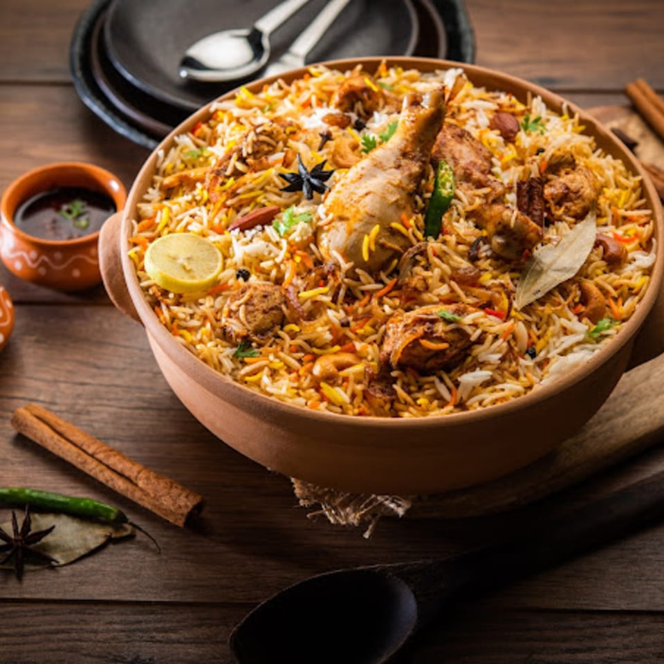 A large bowl of KS Kitchen's chicken biryani with spices and lemon on a wooden table.